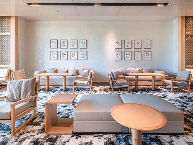 Sala de hotel luminosa con sillas de madera, mesas pequeñas y alfombra gris moderna.