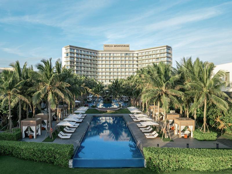 Hotel de lujo con palmeras, sombrillas y una piscina larga y reflejante bajo un cielo azul.