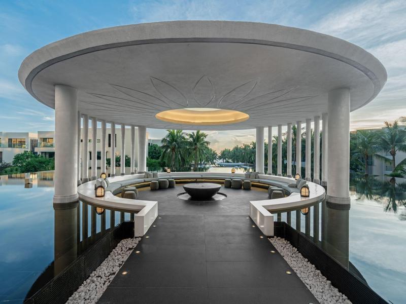 Pabellón redondo con columnas sobre el agua, rodeado de bancos y plantas en complejo hotelero.