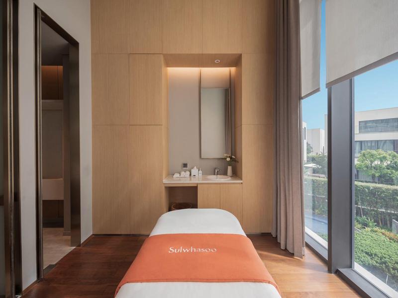 Sala de masajes luminosa con camilla y grandes ventanas con vista a jardines.