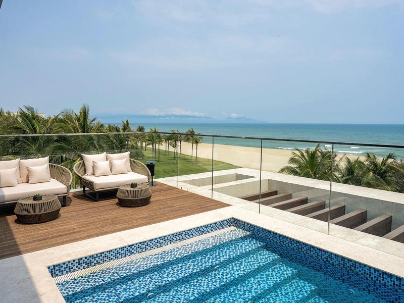 Hotel moderno con piscina y área lounge con vista a palmeras y mar bajo cielo despejado.