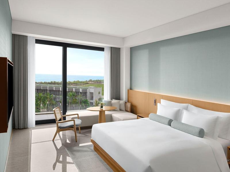 Habitación de hotel luminosa con cama grande, área de estar y balcón con vista a áreas verdes.