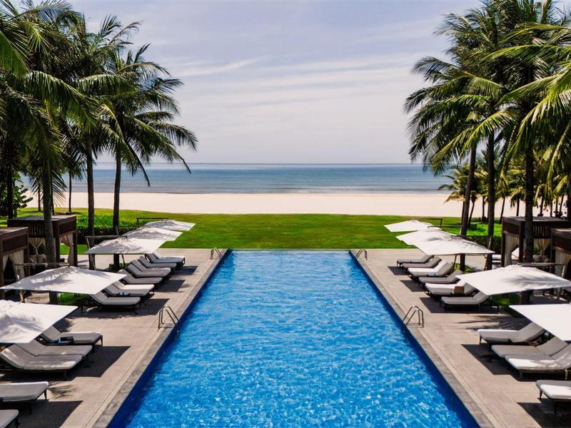 Vista de una piscina con tumbonas y palmeras, con la playa y el mar al fondo.