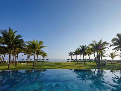 Vista de una piscina con palmeras y el mar al fondo bajo un cielo azul claro.