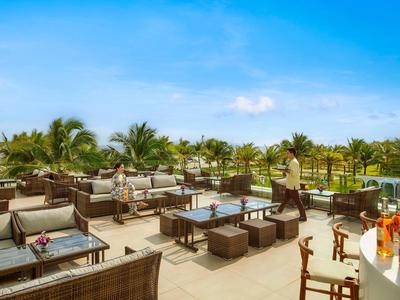 Área exterior de un bar de hotel con asientos, mesas y palmeras bajo un cielo azul.