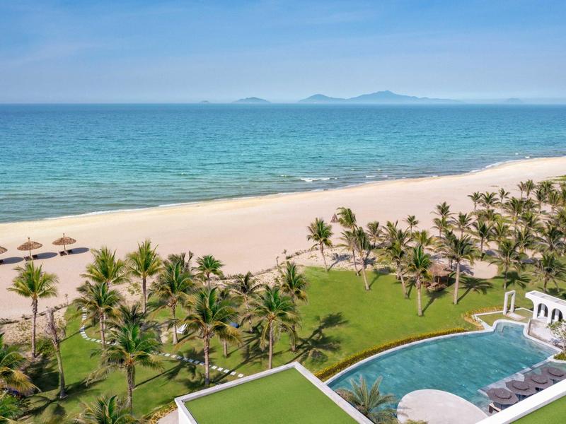 Vista de una playa con palmeras, mar azul y una zona de piscina adyacente de un hotel.
