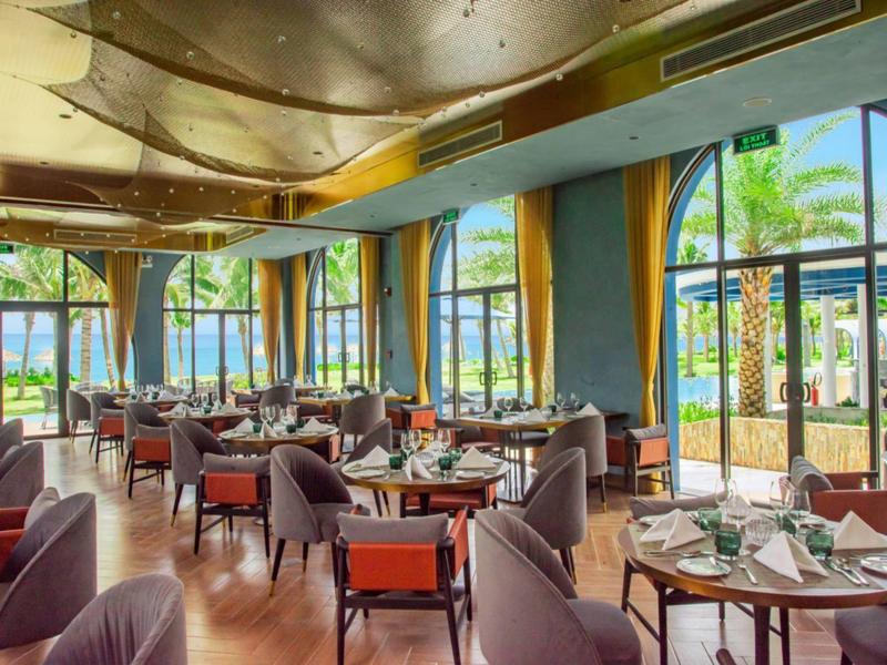 Restaurante de hotel luminoso con grandes ventanas, mesas redondas y decoración moderna.