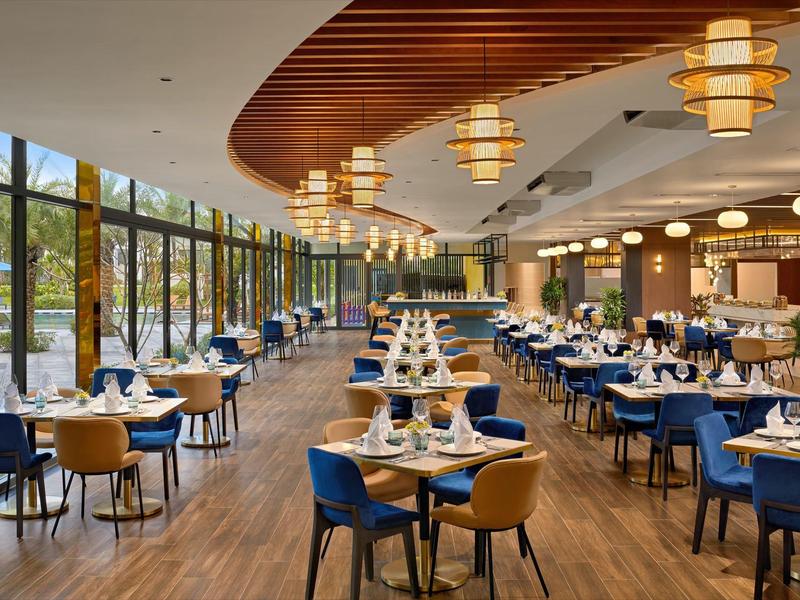 Restaurante moderno con mesas bien dispuestas, sillas azules y amarillas, luces colgantes y grandes ventanas.