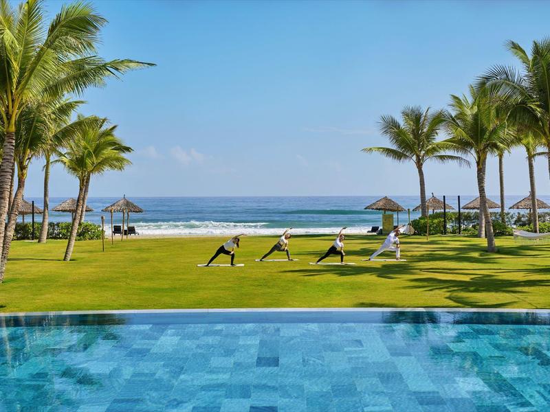 Personas practican yoga en un césped verde junto a una piscina con vista al mar y palmeras.