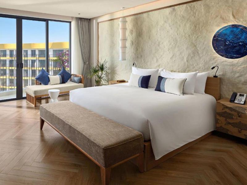 Habitación de hotel luminosa con cama grande, muebles de madera y balcón con vista a un edificio.