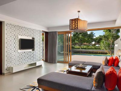 Habitación de hotel luminosa con sofá moderno, televisor grande y vista al jardín a través de puertas de vidrio abiertas.