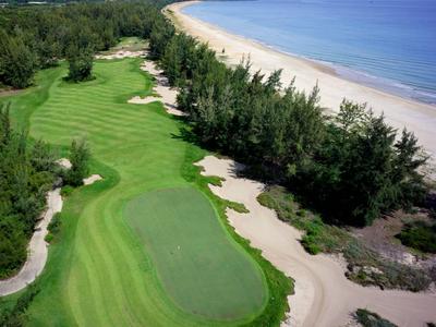 Grüner Golfplatz am Strand mit umliegenden Bäumen und Meer im Hintergrund.