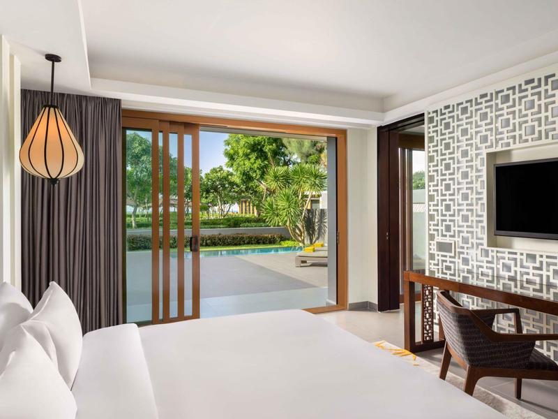 Habitación de hotel luminosa con cama grande, puertas corredizas abiertas y vista a la piscina y jardín