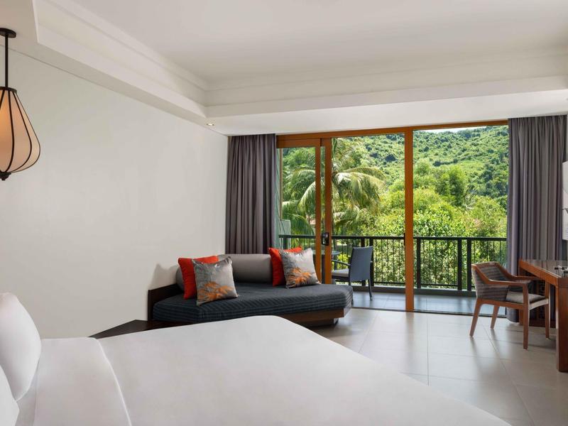 Habitación de hotel moderna con cama, sofá, escritorio y balcón con vista a la naturaleza verde.