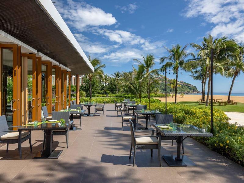 Terraza de hotel con mesas y sillas, rodeada de palmeras y vista a la playa.