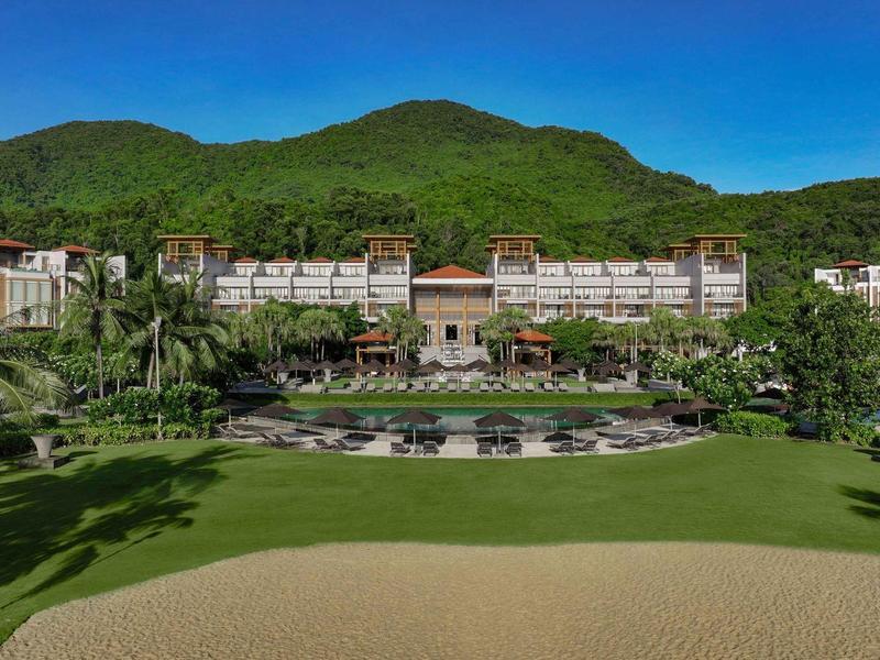 Hotel de lujo frente a montaña verde con gran jardín y zona arenosa.