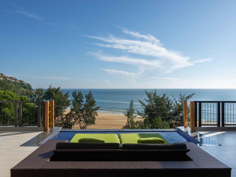 Vista desde una terraza moderna con piscina hacia una playa de arena y el mar bajo un cielo azul.