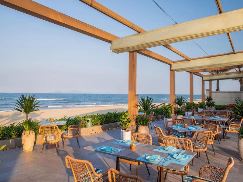 Ristorante aperto sulla spiaggia con tavoli, sedie e vista mare sotto una pergola in legno.