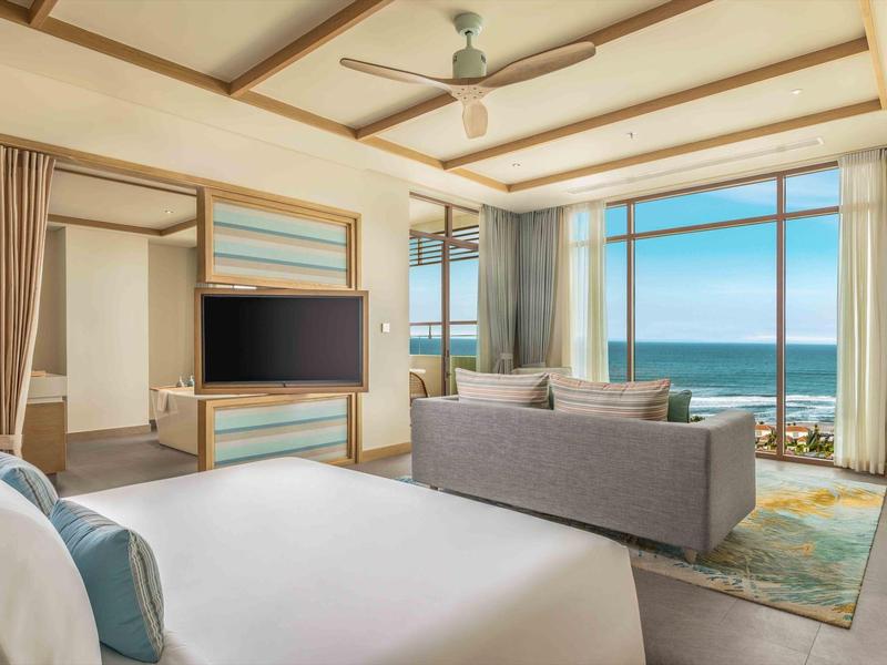 Camera d'hotel moderna con letto, divano, TV e vista mare attraverso grandi finestre.