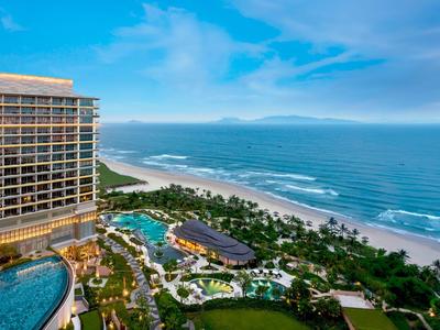 Blick auf ein Hotel am Meer mit Pool, Gartenanlagen und einem weiten Sandstrand bei blauem Himmel.