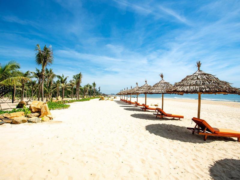 Breed zandstrand met ligstoelen en rieten parasols onder een blauwe lucht aan zee.