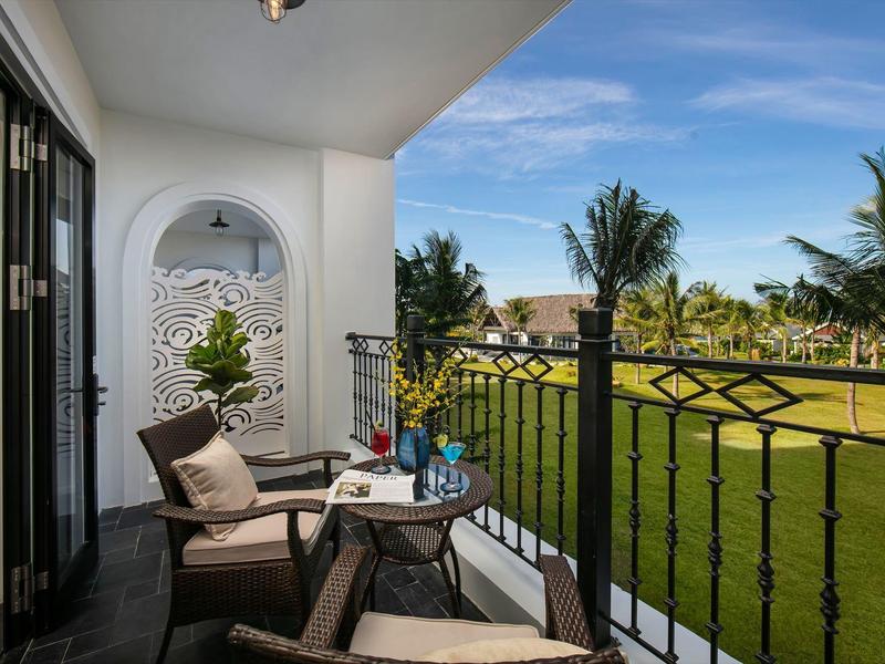 Balkon met zitplaatsen en uitzicht op verzorgde tuin en palmbomen onder blauwe lucht.