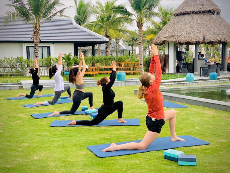 Mensen doen yoga op matten in een hotel tuin met palmbomen en zwembad.