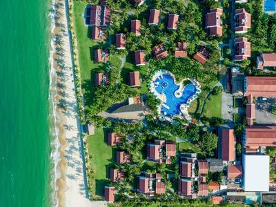 Luchtfoto van een resort met huisjes en een groot zwembad bij het strand.