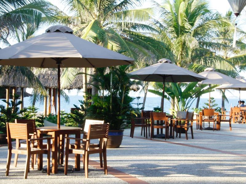 Buitengebied met houten tafels, stoelen en parasols omringd door palmbomen bij het water.