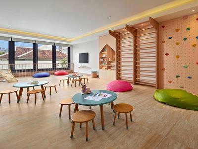 Sala giochi luminosa per bambini con parete arrampicata, piccoli tavoli, sgabelli e grandi finestre con vista.