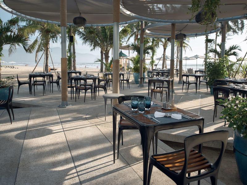 Area salotto aperta con sedie e tavoli in legno sotto una tettoia vicino alla spiaggia.