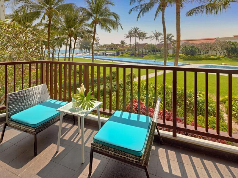 Balcone con due sedie e tavolo che si affaccia su piscina tropicale e palme.