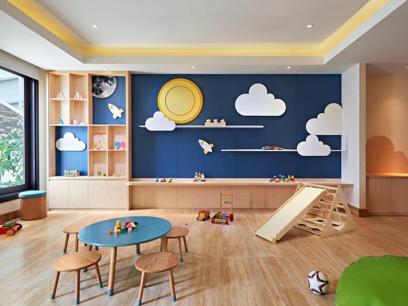 Sala giochi luminosa per bambini con giocattoli in legno, tavoli e scivolo davanti a una parete blu con decorazioni di nuvole e sole.