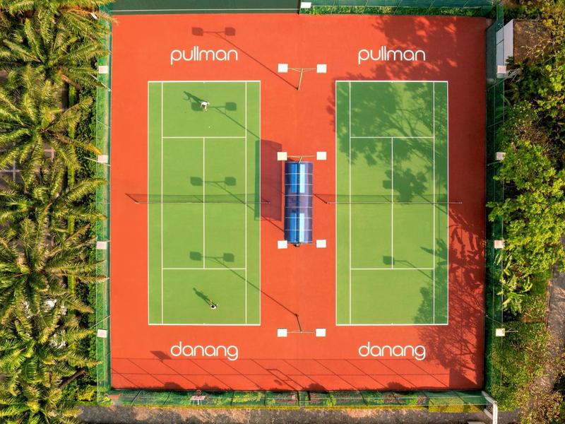 Due campi da tennis verdi con superficie rossa, circondati da alberi e palme.