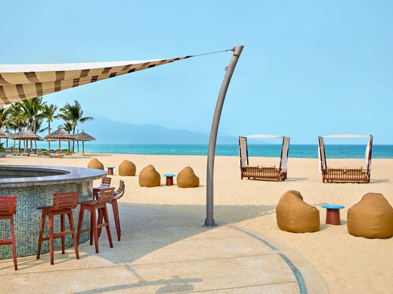 Bar sulla spiaggia con posti a sedere, lettini e spiaggia sabbiosa davanti al mare blu e cielo sereno.