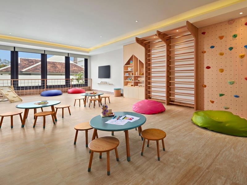 Sala giochi luminosa per bambini con parete arrampicata, piccoli tavoli, sgabelli e grandi finestre con vista.