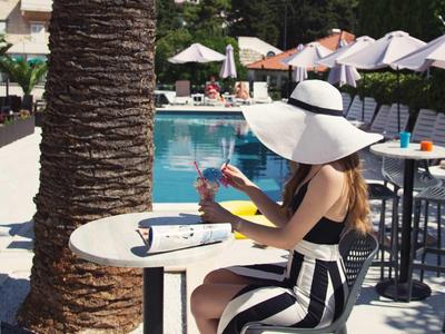 Frau in gestreiftem Kleid mit großem Hut sitzt am Pool an einem Tisch.