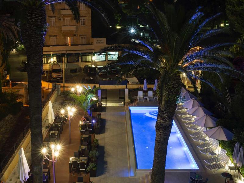 Nächtliche Terrasse mit blau beleuchtetem Pool, Palmen und gedeckten Tischen in warmem Licht.