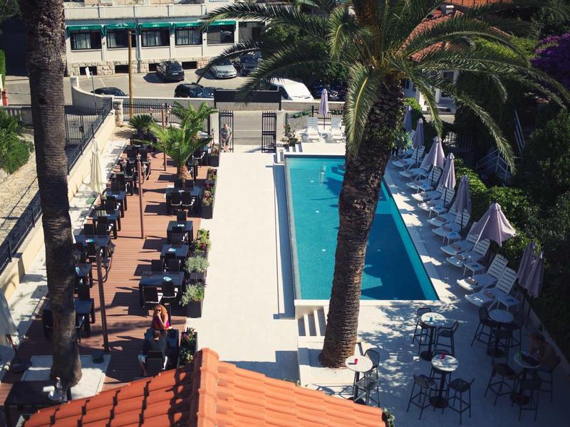 Hotelterrasse mit Pool, Liegestühlen und Palmen, umgeben von Gebäuden und Sonnenschirmen.