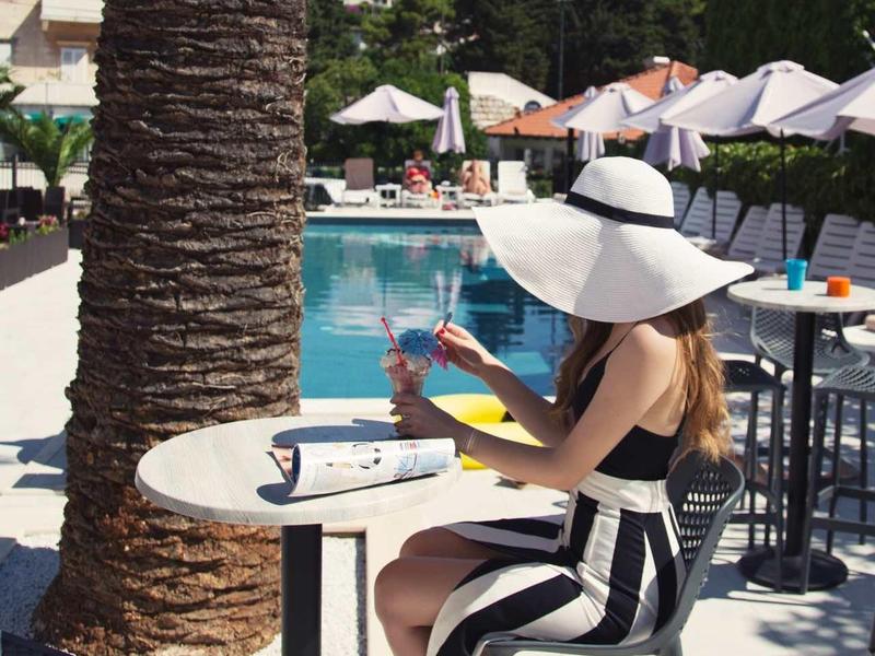 Frau mit großem Hut sitzt an einem Tisch, Pool und Sonnenschirme im Hintergrund.