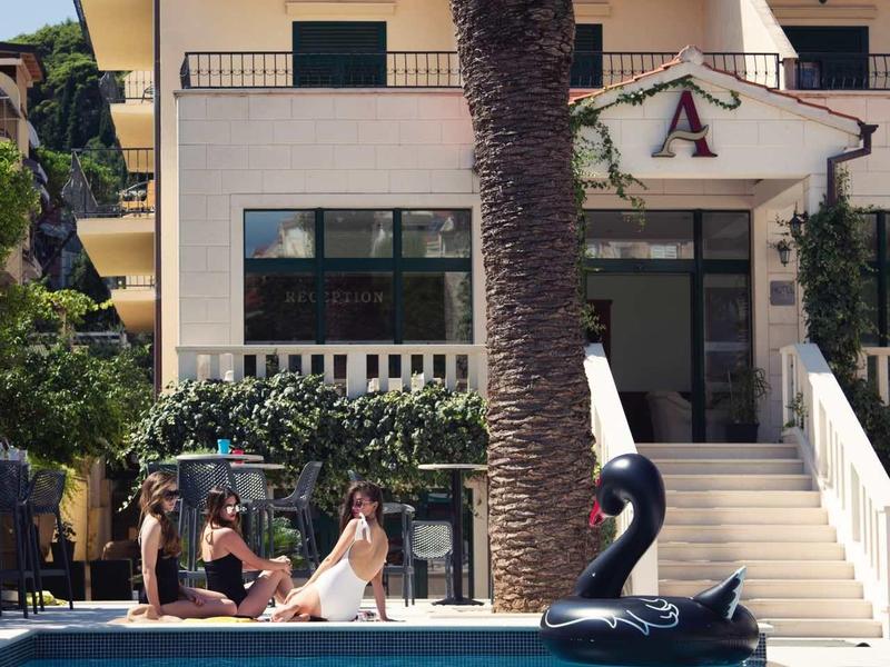 Pool mit klarem Wasser, Palme im Vordergrund, Menschen entspannen und aufblasbarer Schwanschwarz.