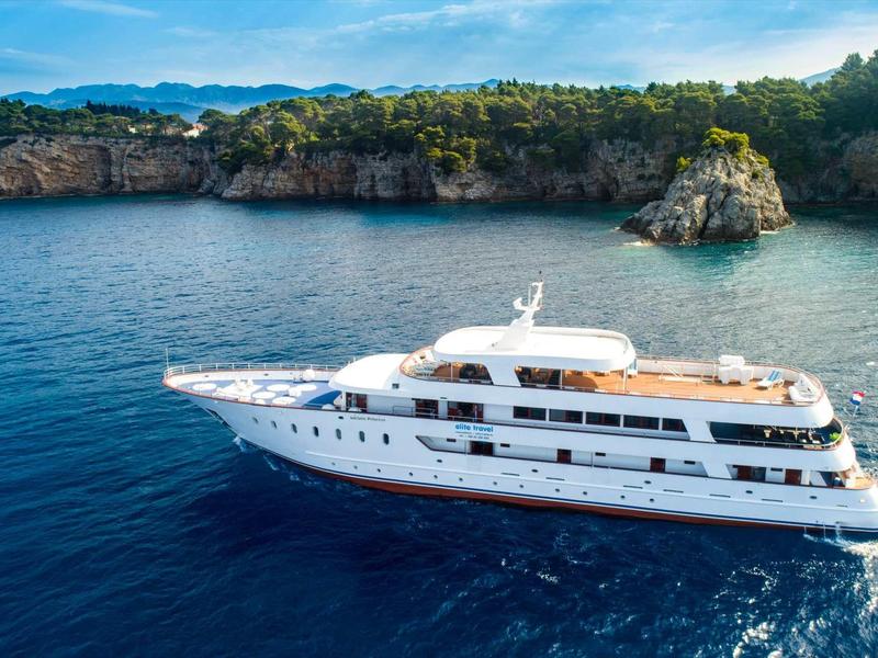 Luxury white yacht sailing in clear blue water near rocky coast with green vegetation.