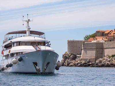 Ein großes weißes Kreuzfahrtschiff fährt nah an einer steinernen Küstenmauer mit Gebäuden vorbei.