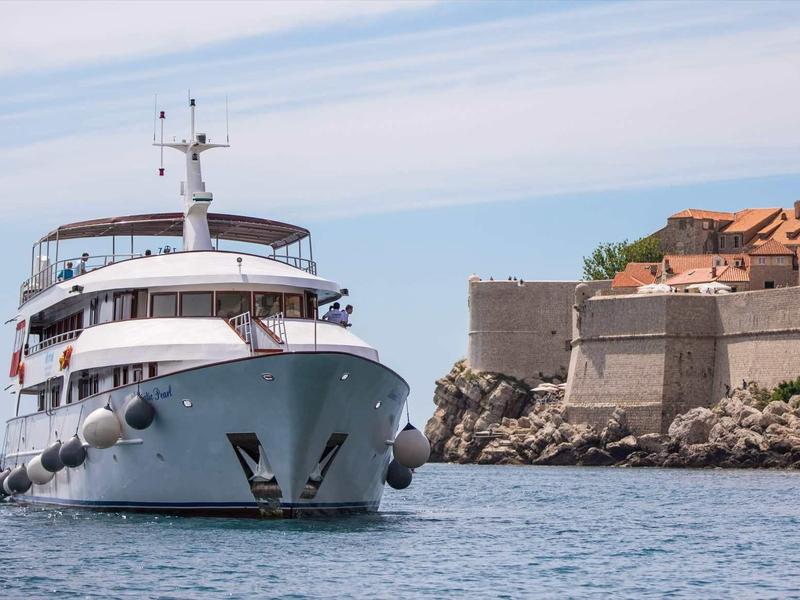 Ein großes weißes Kreuzfahrtschiff fährt nah an einer steinernen Küstenmauer mit Gebäuden vorbei.