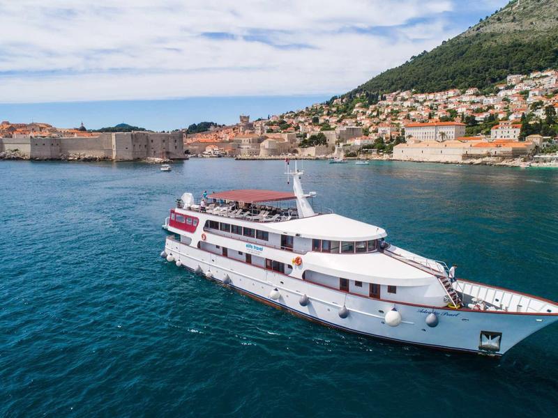Ein weißes Passagierschiff auf blauem Meer vor einer Küstenstadt mit Häusern und Hügeln.
