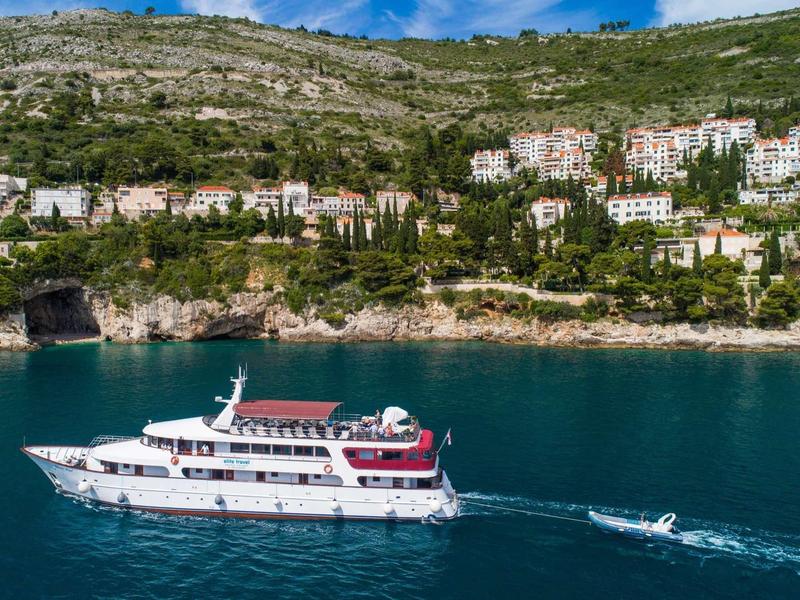 Weißes Boot fährt auf klarem blauem Wasser vor bewaldetem Hang mit Häusern und blauem Himmel.