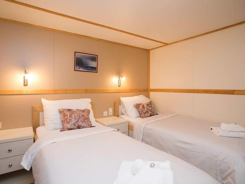 Kleines Zimmer mit zwei Einzelbetten, weißen Bettwäsche, Nachttisch und warmem Licht an der Wand.