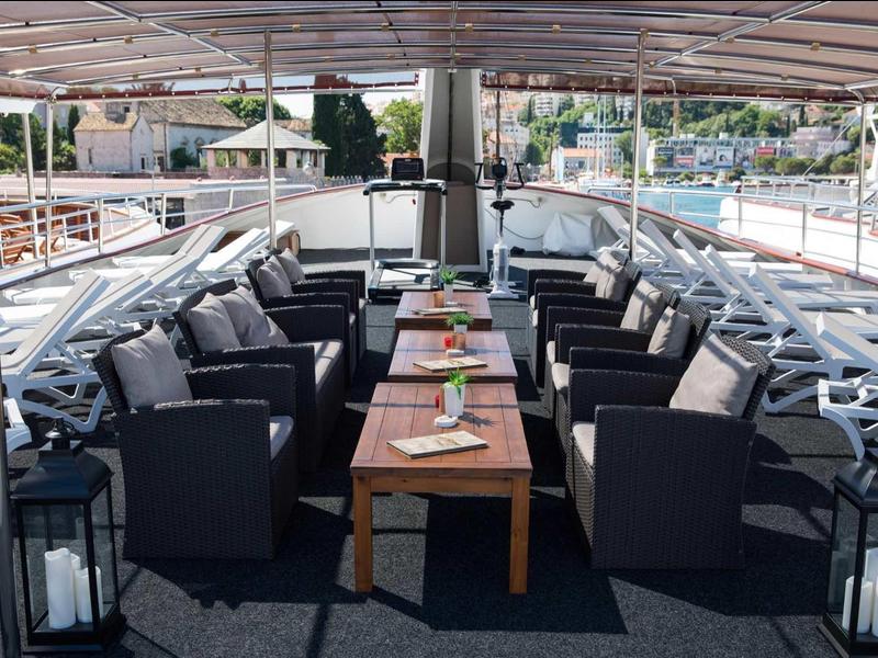 Großer, gedeckter Sitzbereich auf einem Boot mit grauen Polstersesseln und Tisch in der Mitte.