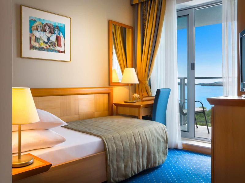 Bright hotel room with single bed, desk, lamp, and balcony overlooking the sea.