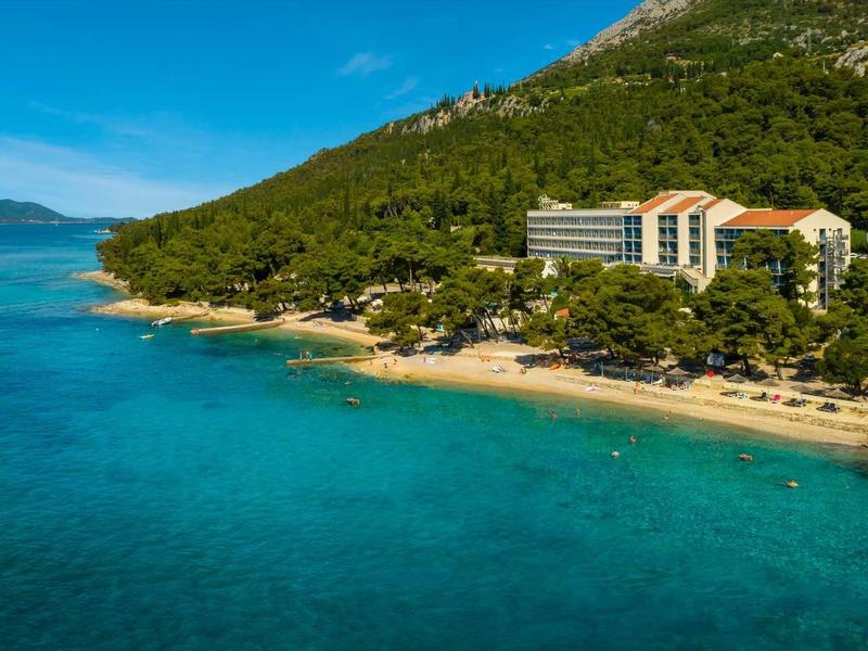 Hotel on green hillside by clear blue sea with sandy beach and forested surroundings.
